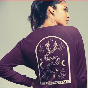 May include: A maroon long-sleeve sweatshirt with a white graphic of two snakes, stars, and crescent moons inside an arch. The text "FIND YOUR OWN PATH" is at the bottom of the graphic.