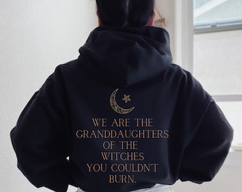 May include: Black hooded sweatshirt with a gold crescent moon and star graphic on the back. The text "We are the granddaughters of the witches you couldn't burn." is printed below the graphic.