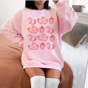May include: A pink sweatshirt with a graphic print of red strawberries. The sweatshirt has long sleeves and a crew neck.