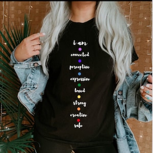 May include: Black t-shirt with the text "i am connected perceptive expressive loved strong creative safe" printed vertically down the center. Each word has a colored dot next to it, forming a rainbow.