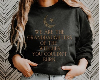 Może przedstawiać: Czarna bluza z nadrukiem złotego półksiężyca i gwiazdy. Na przodzie znajduje się napis "We are the granddaughters of the witches you couldn't burn."