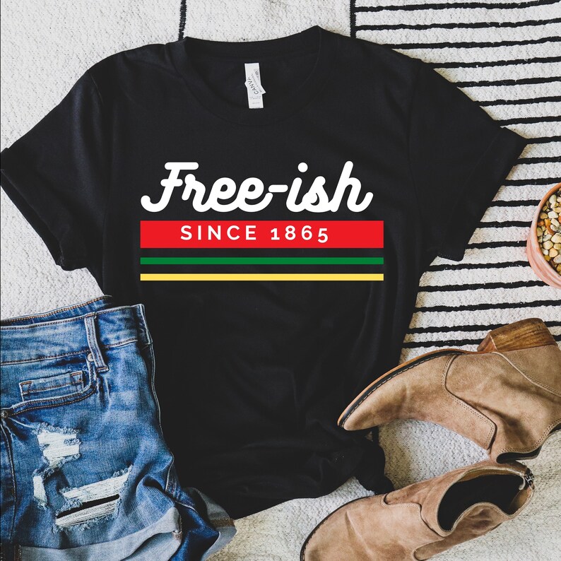 May include: Black t-shirt with the text "Free-ish SINCE 1865" in white, red, green, and yellow. The shirt is displayed with denim shorts and brown suede boots on a striped rug.