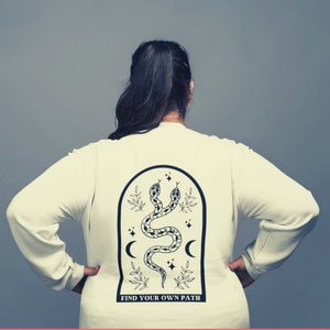 May include: Cream-colored long-sleeve shirt with a black graphic on the back. The graphic features two snakes, crescent moons, stars, and the text "FIND YOUR OWN PATH" within an arched frame.