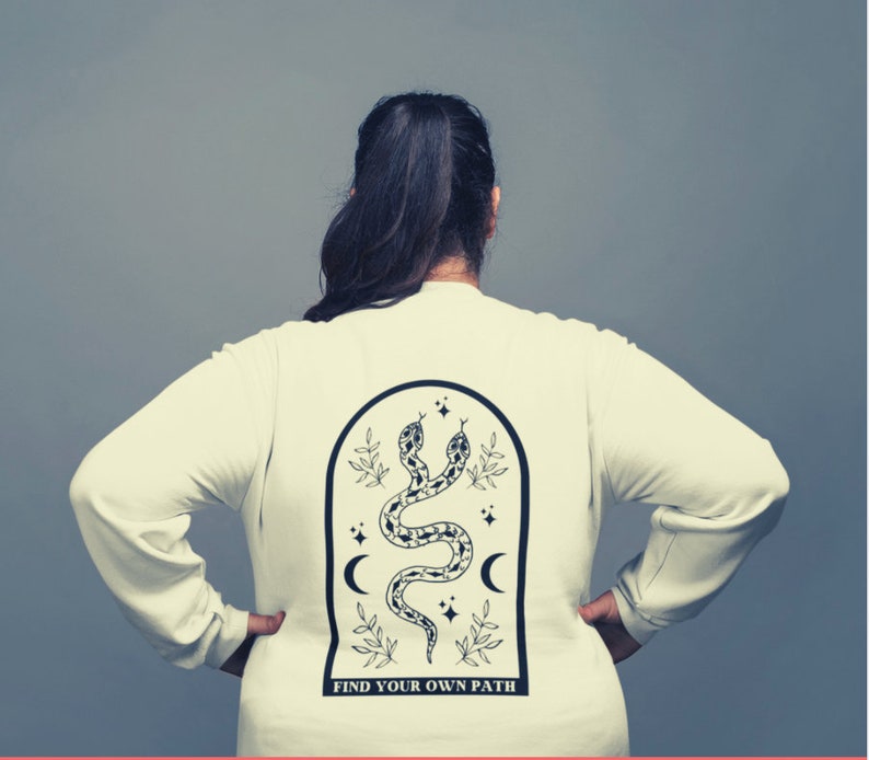 May include: Cream-colored long-sleeve shirt with a black graphic on the back. The graphic features two snakes, crescent moons, stars, and the text "FIND YOUR OWN PATH" within an arched frame.