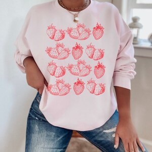 May include: A pink sweatshirt with a graphic print of red strawberries. The strawberries are arranged in rows and are outlined in black.