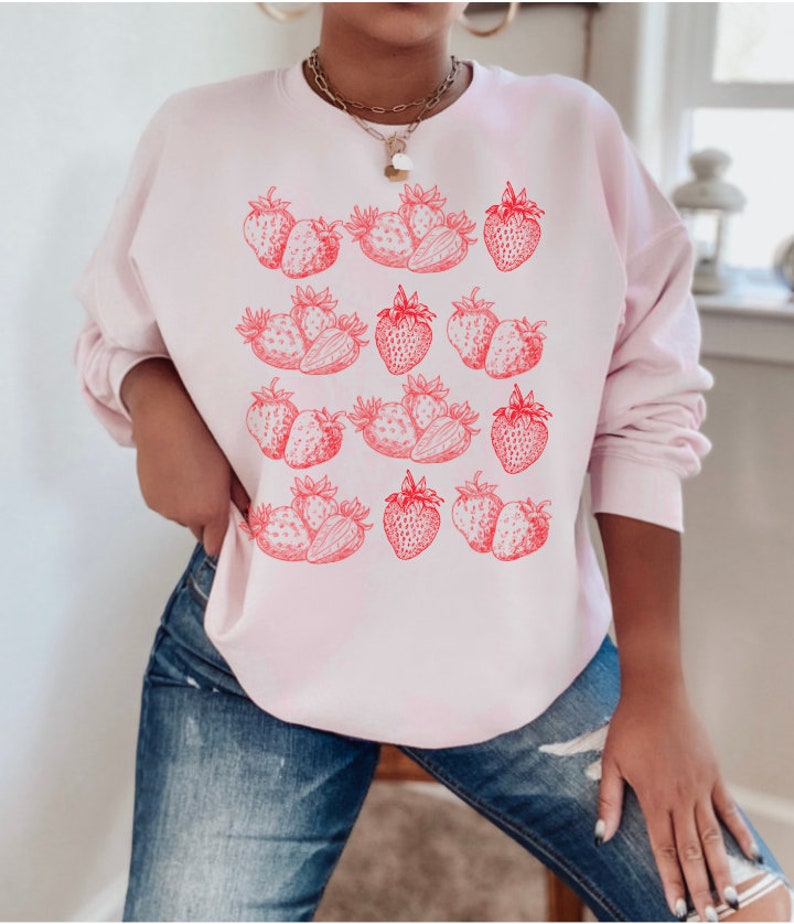 May include: A pink sweatshirt with a graphic print of red strawberries. The strawberries are arranged in rows and are outlined in black.