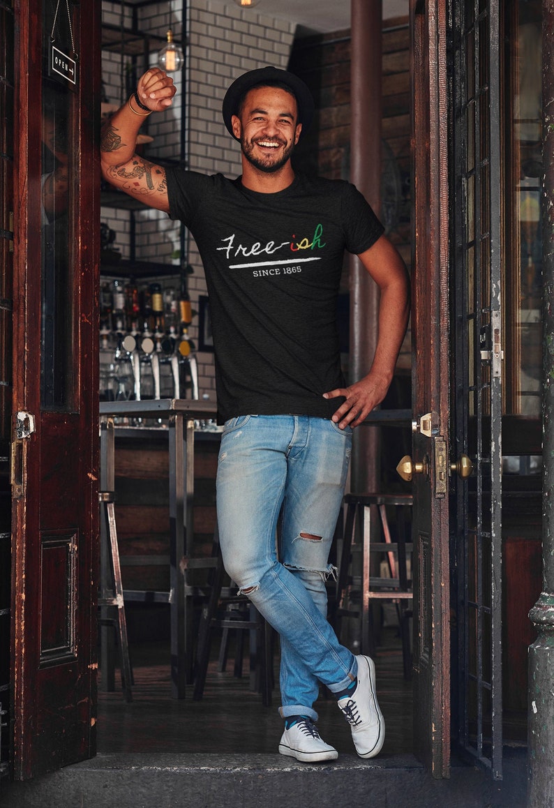 Pode incluir: Camiseta preta com o texto "Free-ish SINCE 1865" em branco, verde e vermelho. A camiseta &eacute; usada com jeans azul claro e t&ecirc;nis brancos. A pessoa est&aacute; encostada em uma moldura de porta de madeira.