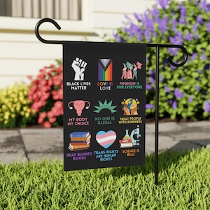 May include: A black and white garden flag with a variety of colourful illustrations and text. The text includes "Black Lives Matter", "Love is Love", "Feminism is for Everyone", "My Body My Choice", "No One is Illegal", "Treat People with Kindness", "Read Banned Books", "Trans Rights are Human Rights", and "Science is Real".