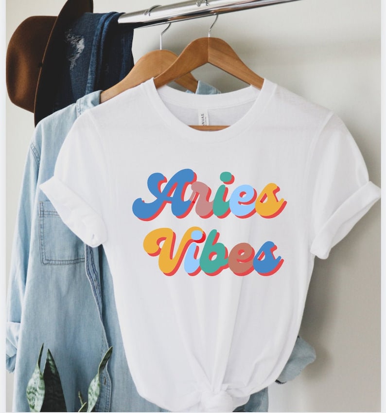 Op de afbeelding: Wit T-shirt met een kleurrijke retro-stijl grafische afbeelding met de tekst "Aries Vibes".