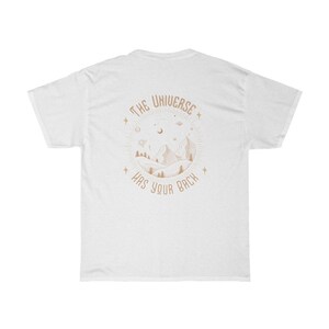 Pode incluir: Camiseta branca com um gr&aacute;fico circular nas costas. O gr&aacute;fico apresenta uma cordilheira, &aacute;rvores e elementos celestes em marrom claro. O texto "THE UNIVERSE HAS YOUR BACK" est&aacute; arqueado ao redor do design.