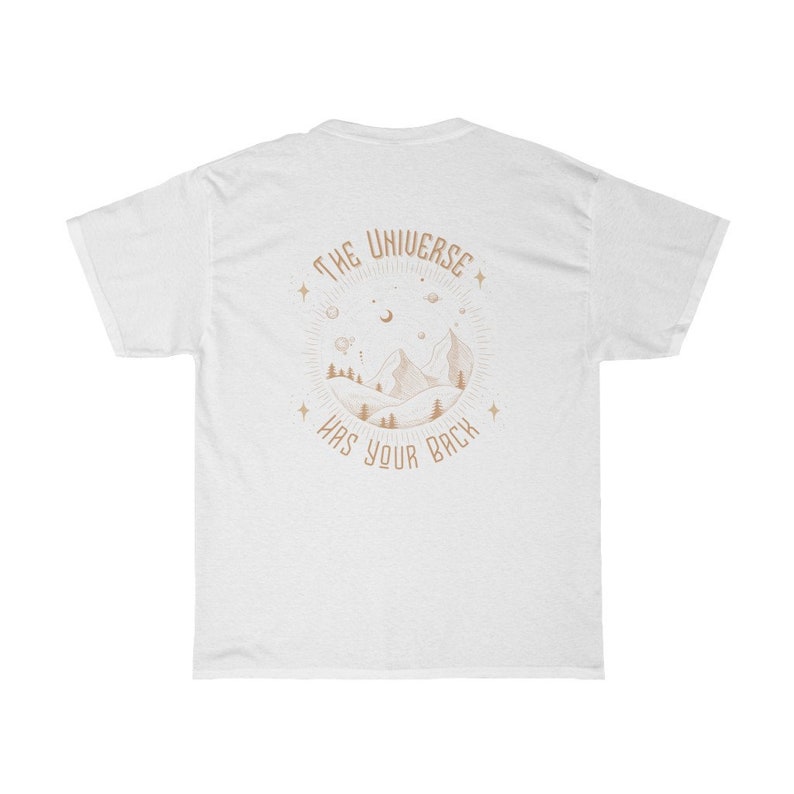 Pode incluir: Camiseta branca com um gr&aacute;fico circular nas costas. O gr&aacute;fico apresenta uma cordilheira, &aacute;rvores e elementos celestes em marrom claro. O texto "THE UNIVERSE HAS YOUR BACK" est&aacute; arqueado ao redor do design.