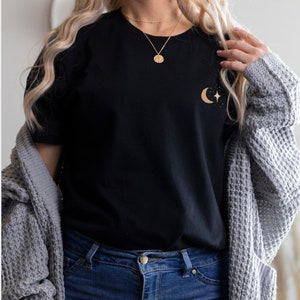 Pode incluir: Camiseta preta com um design dourado de lua crescente e estrela. A camisa &eacute; combinada com jeans azuis e um cardigan cinza. Um colar de ouro tamb&eacute;m &eacute; vis&iacute;vel.