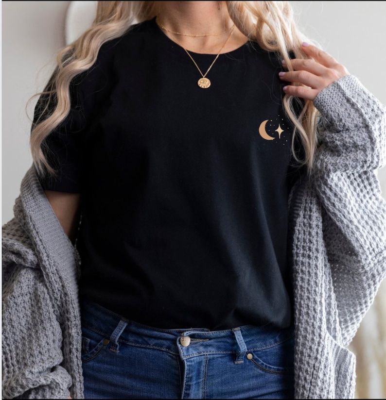 Pode incluir: Camiseta preta com um design dourado de lua crescente e estrela. A camisa &eacute; combinada com jeans azuis e um cardigan cinza. Um colar de ouro tamb&eacute;m &eacute; vis&iacute;vel.