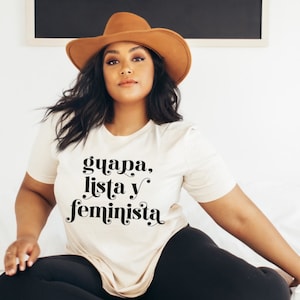 May include: A beige t-shirt with the words "guapa, lista y feminista" in black script. The person in the image is wearing a brown hat and black pants. The shirt is a casual, everyday wear item.