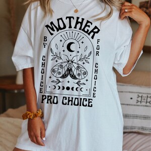 Pu&ograve; includere: Maglietta bianca con un design grafico nero che presenta una falena, una luna crescente e stelle all'interno di un arco. Il testo "Mother by Choice for Choice Pro Choice" &egrave; stampato sotto il grafico.