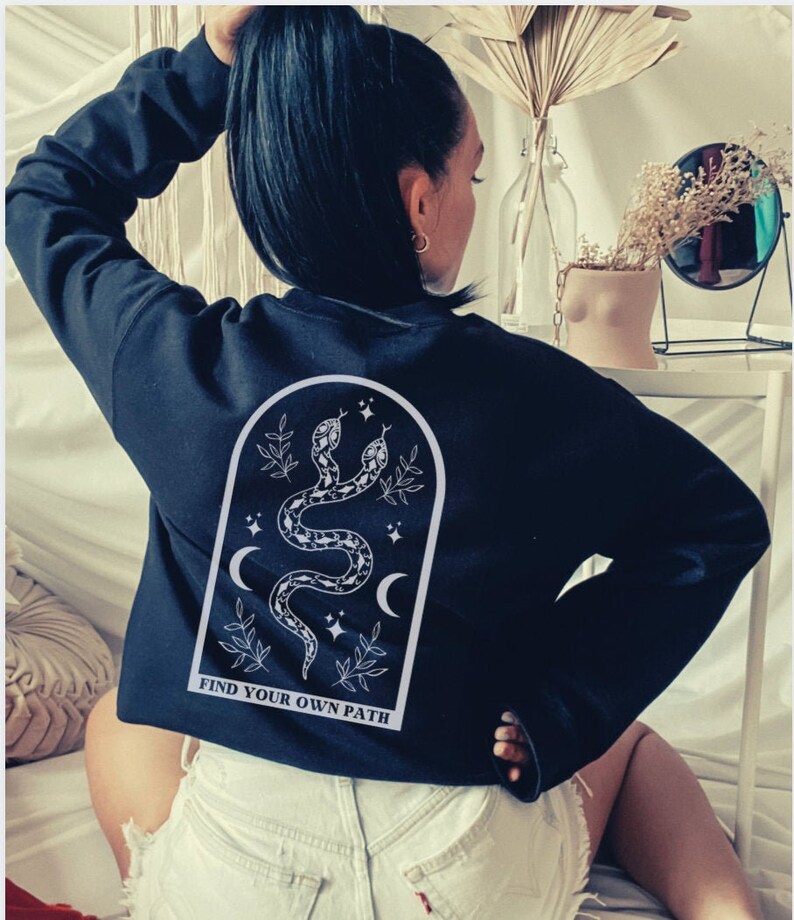 May include: Black sweatshirt with a white graphic on the back featuring two snakes, crescent moons, stars, and the text "FIND YOUR OWN PATH". The sweatshirt is a crew neck style.