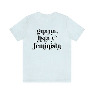 May include: A light blue t-shirt with the words "guapa, lista y feminista" in black script. The text is centered on the front of the shirt. The shirt has short sleeves and a crew neck.