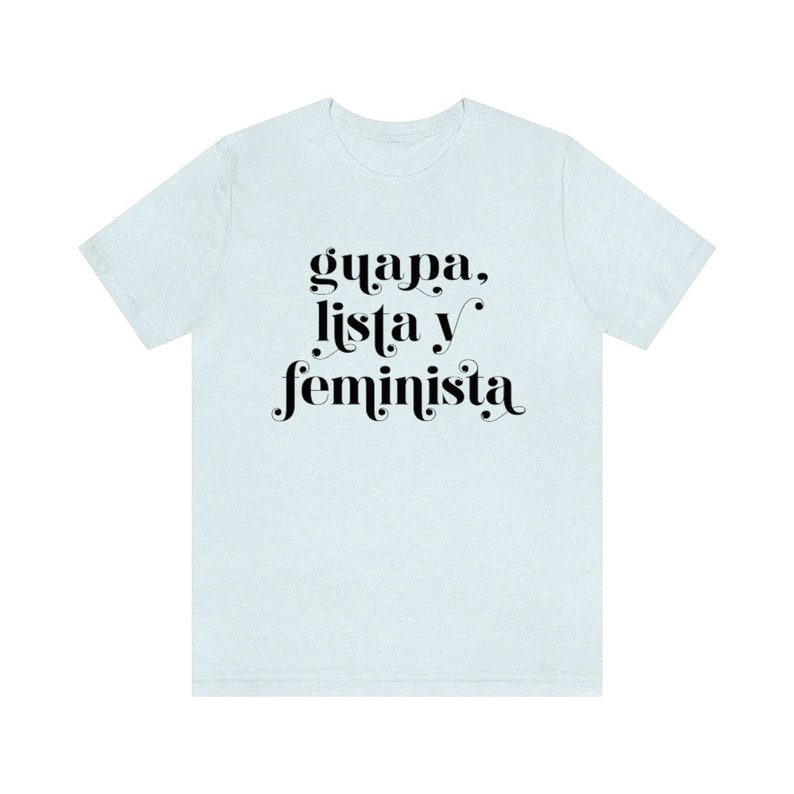 May include: A light blue t-shirt with the words "guapa, lista y feminista" in black script. The text is centered on the front of the shirt. The shirt has short sleeves and a crew neck.