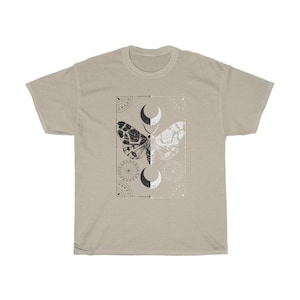 K&ouml;nnte beinhalten: Ein beiges T-Shirt mit einem schwarzen und wei&szlig;en Mottivendesign. Der Falter befindet sich in der Mitte eines quadratischen Rahmens mit geometrischen Mustern und Mondsicheln.