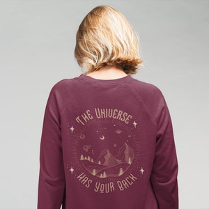 Puede incluir: Sudadera color burdeos con un estampado gr&aacute;fico de una cordillera y estrellas con el texto "The Universe Has Your Back" en color dorado.
