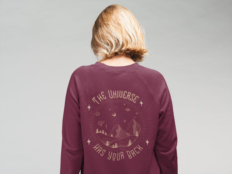 Puede incluir: Sudadera color burdeos con un estampado gr&aacute;fico de una cordillera y estrellas con el texto "The Universe Has Your Back" en color dorado.