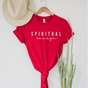 May include: A red t-shirt with the words "SPIRITUAL savage" printed in white. The shirt is hanging on a wooden hanger, with a beige hat visible in the background. The shirt is knotted at the bottom.