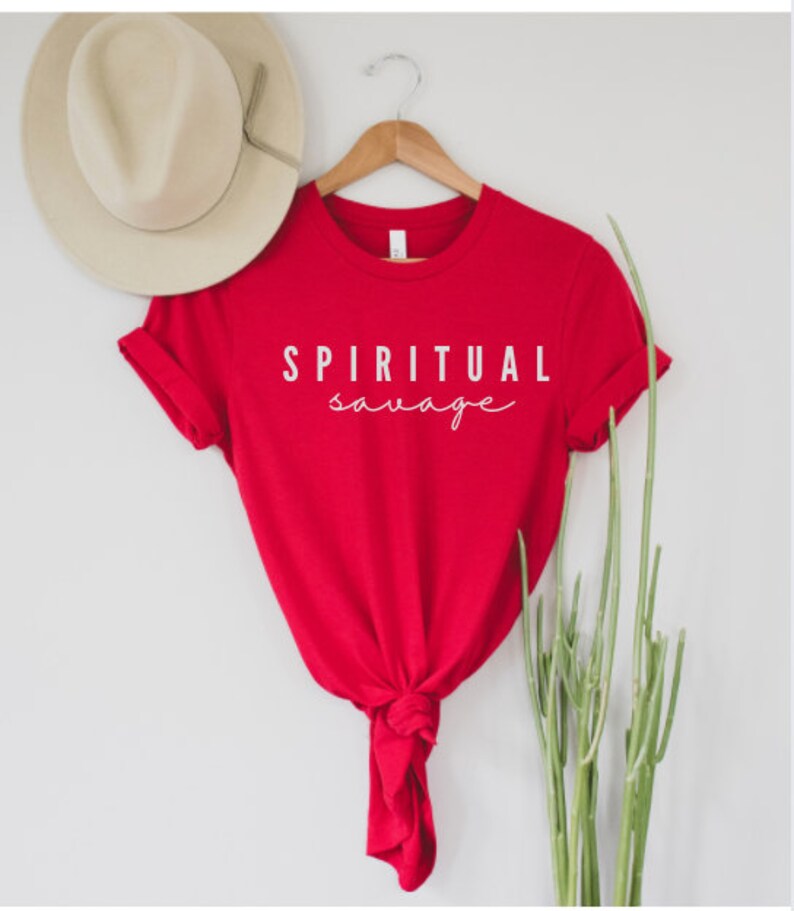 May include: A red t-shirt with the words "SPIRITUAL savage" printed in white. The shirt is hanging on a wooden hanger, with a beige hat visible in the background. The shirt is knotted at the bottom.