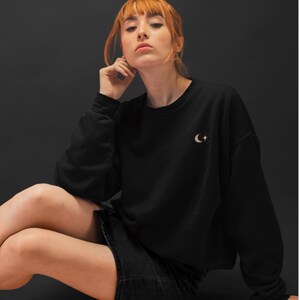 May include: A black sweatshirt with a crescent moon and stars design in gold embroidery. The model is wearing a black skirt and has red hair.