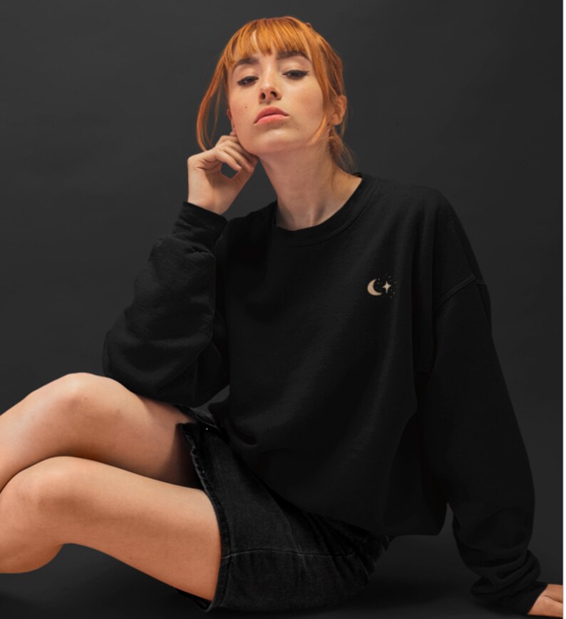 May include: A black sweatshirt with a crescent moon and stars design in gold embroidery. The model is wearing a black skirt and has red hair.