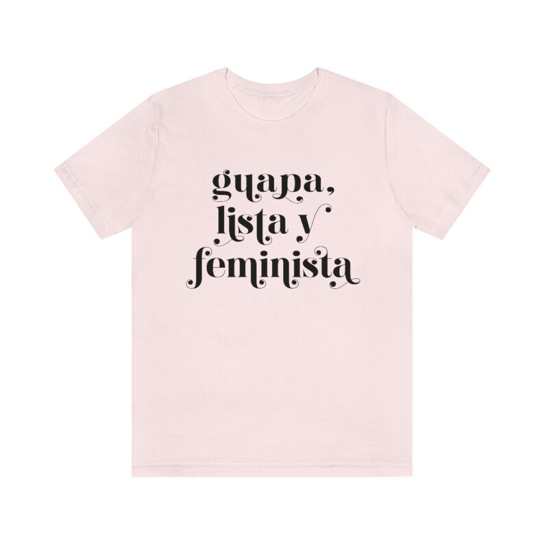 May include: A light pink t-shirt with the words "guapa, lista y feminista" printed in black, stylized font. The shirt is a short-sleeved crew neck style.