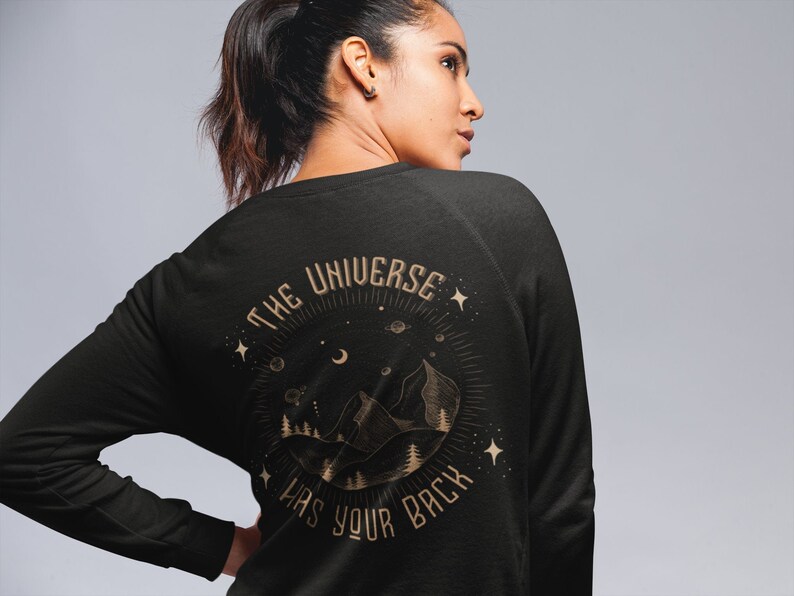 May include: Black long-sleeve sweatshirt with a gold graphic print of a mountain range, stars, and a crescent moon. The text "The Universe Has Your Back" is printed in a circle around the graphic.