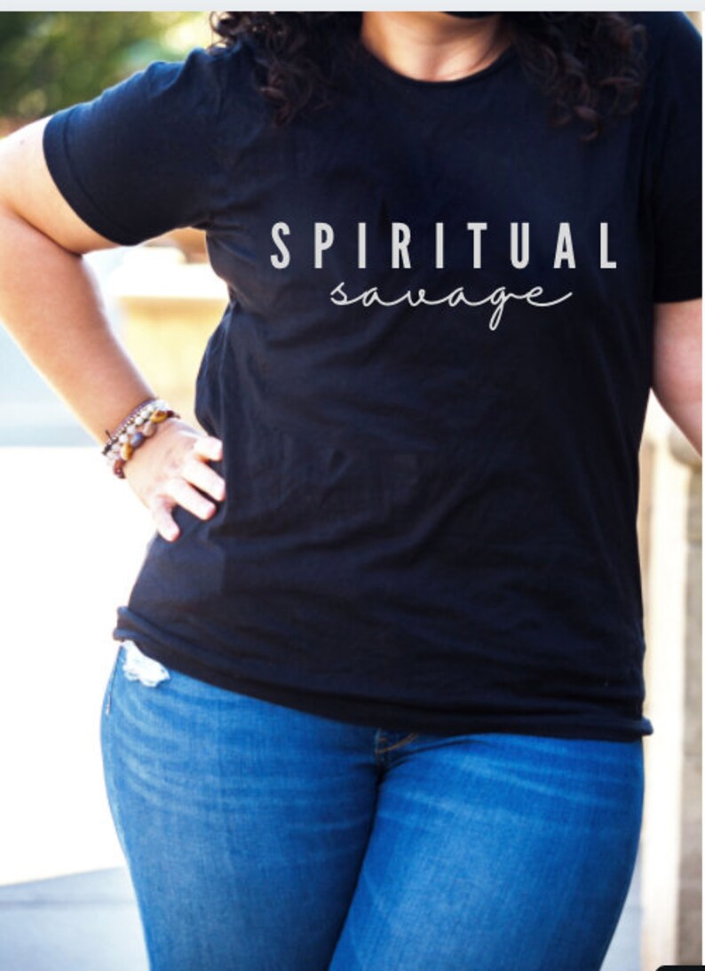 May include: Black t-shirt with the words "SPIRITUAL savage" printed in white. The model is wearing blue jeans and a bracelet. The shirt is a crew neck and short-sleeved.