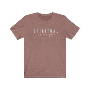 May include: A dusty rose-colored t-shirt with the words "SPIRITUAL savage" printed in a light gray color. The word "spiritual" is in a sans-serif font, and the word "savage" is in a cursive font.