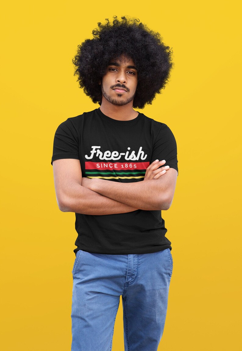 May include: Black t-shirt with the text "Free-ish Since 1865" in white script above a red, green, and yellow stripe design. The shirt is worn by a person with an afro and blue jeans against a yellow background.