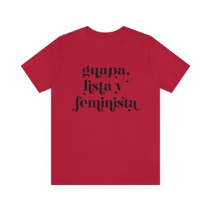 May include: A red t-shirt with the words "guapa, lista y feminista" in black script. The text is centered on the front of the shirt. The shirt has short sleeves and a crew neck.