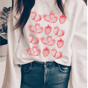 May include: White sweatshirt with a red strawberry print. The strawberries are arranged in a pattern on the front of the sweatshirt.