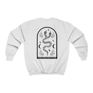 May include: White sweatshirt featuring a black graphic of two intertwined snakes within an arched frame. The design includes crescent moons, stars, and the text "FIND YOUR OWN PATH" at the bottom. The sweatshirt has long sleeves and a crew neck.
