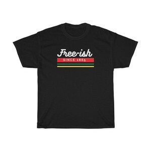 May include: Black t-shirt with the text "Free-ish Since 1865" in white script above a horizontal stripe design in red, green, and yellow. The shirt is a casual wear item.