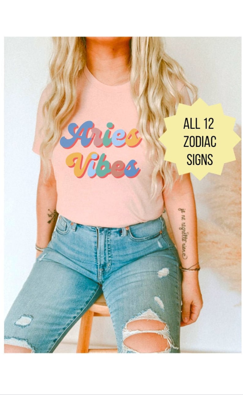 Op de afbeelding: Een roze T-shirt met de tekst "Aries Vibes" in kleurrijke retro letters. De tekst "ALL 12 ZODIAC SIGNS" staat in een gele stervorm.