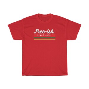 May include: A red t-shirt with the words "Free-ish" in white script above a horizontal stripe design in red, green, and yellow, with "SINCE 1865" below. The shirt is made of a soft material.