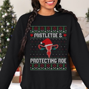 Peut inclure: Sweat-shirt noir avec un motif festif. Le motif comprend un utérus portant un bonnet de Père Noël, avec les mots "Mistletoe & Protecting Roe" en blanc et vert. Le motif est de style pull de Noël.
