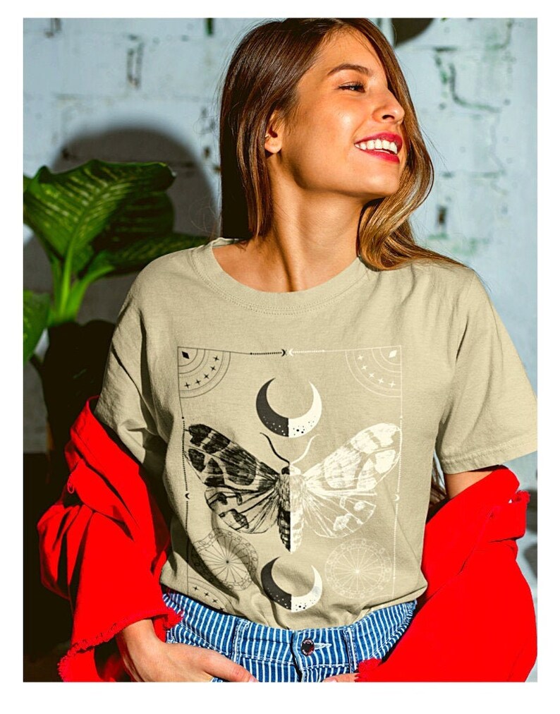 K&ouml;nnte beinhalten: Ein hellbeiges T-Shirt mit einem grafischen Druck eines Nachtfalters mit ausgebreiteten Fl&uuml;geln und einem Halbmond-Design. Der Nachtfalter ist schwarzwei&szlig; und der Mond ist schwarzwei&szlig; mit einer Halbmondform.