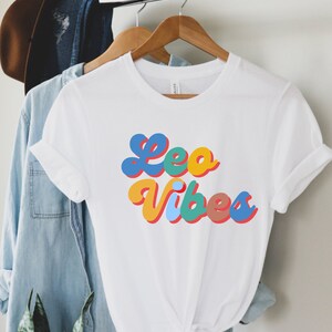 Op de afbeelding: Wit T-shirt met een kleurrijke retro-stijl grafische afbeelding met de tekst "Leo Vibes".