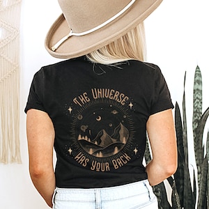 Pode incluir: Camiseta preta com um gr&aacute;fico circular com montanhas, &aacute;rvores, estrelas e o texto "THE UNIVERSE HAS YOUR BACK" em dourado. A camiseta &eacute; de gola redonda.