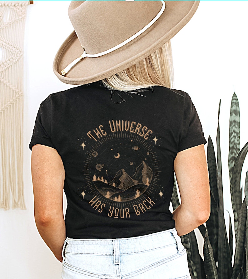 Pode incluir: Camiseta preta com um gr&aacute;fico circular com montanhas, &aacute;rvores, estrelas e o texto "THE UNIVERSE HAS YOUR BACK" em dourado. A camiseta &eacute; de gola redonda.