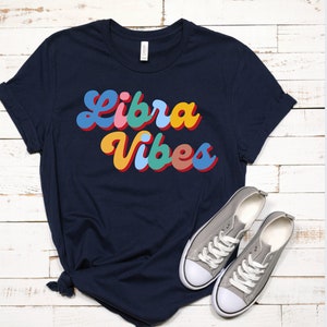 Op de afbeelding: Een marineblauw t-shirt met de tekst "Libra Vibes" in kleurrijke retro letters.
