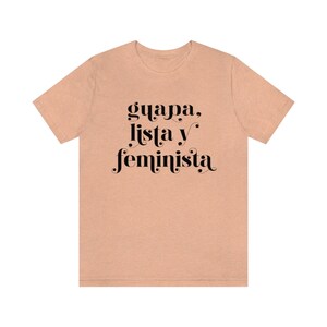 May include: A peach-colored t-shirt with the words "guapa, lista y feminista" printed in black script. The text is centered on the front of the shirt, with the words arranged vertically. The shirt has short sleeves and a crew neck.