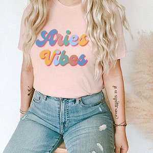 Op de afbeelding: Een roze T-shirt met de tekst "Aries Vibes" in kleurrijke retro letters. Het shirt wordt gedragen door een persoon met lang blond haar en een blauwe jeans.