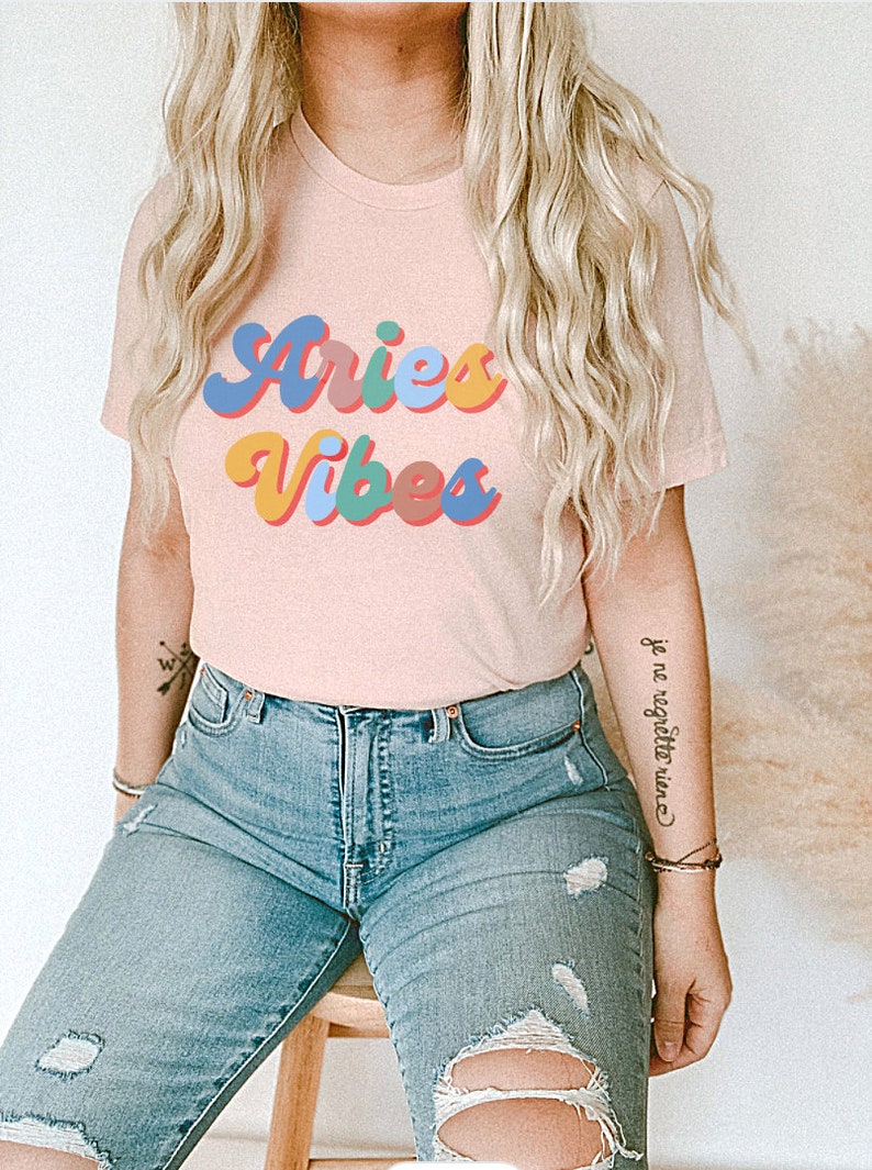 Op de afbeelding: Een roze T-shirt met de tekst "Aries Vibes" in kleurrijke retro letters. Het shirt wordt gedragen door een persoon met lang blond haar en een blauwe jeans.