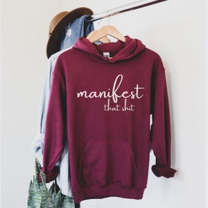 May include: A maroon hoodie with the words "manifest that shit" in white script. The hoodie has a front pocket and a hood. A brown hat and a blue denim shirt hang behind the hoodie on a clothing rack.